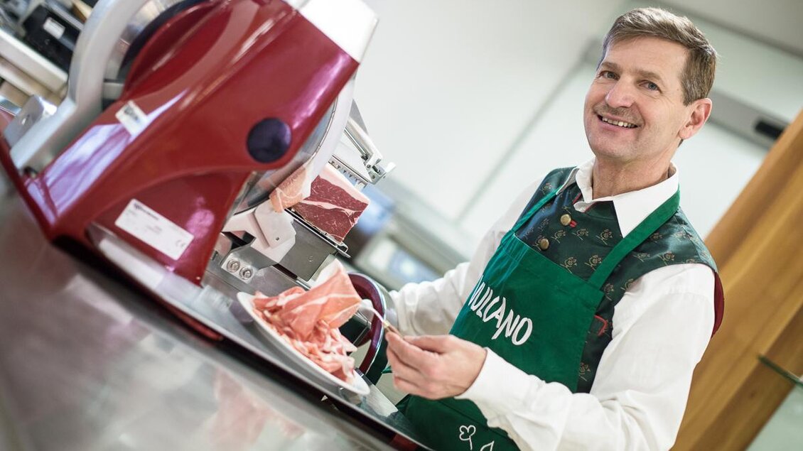 Ein freundlicher Metzger mit einem grünen Schürze steht an einer Aufschnittmaschine. Er hält einen Teller mit frisch geschnittenem Fleisch in der Hand. | © Vulcano Schinkenmanufaktur