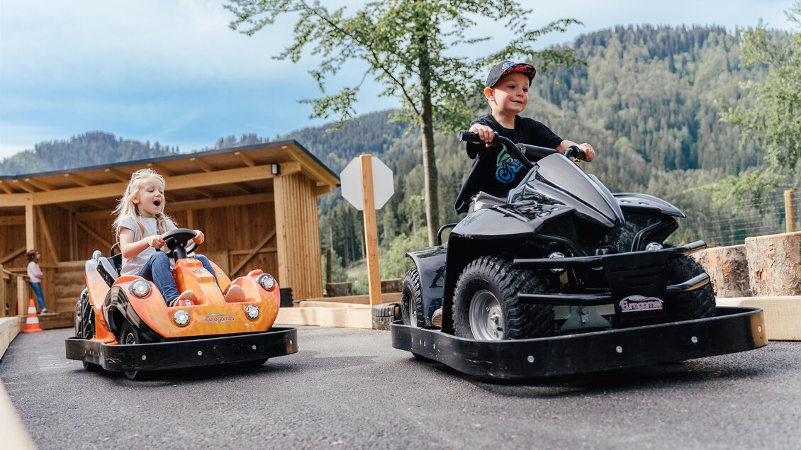 Zwei Kinder fahren auf einer Kartbahn. Der Junge fährt ein schwarzes Fahrzeug, während das Mädchen in einem orangefarbenen Kart sitzt. | © Waldpark Hochreiter