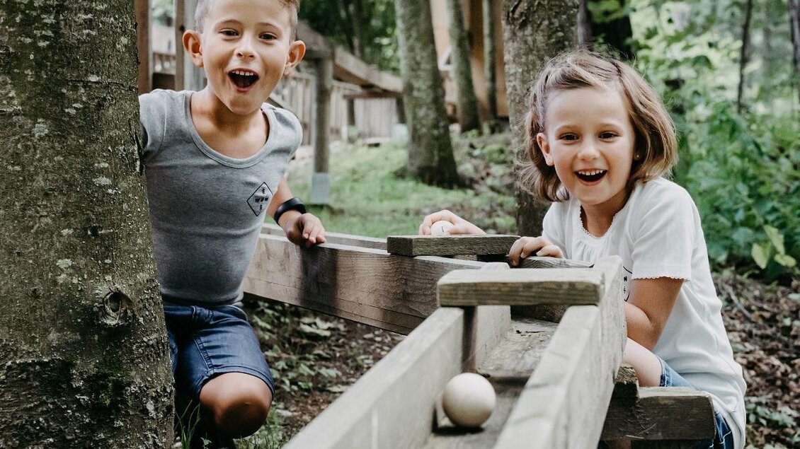 Zwei fröhliche Kinder spielen im Freien. Sie lächeln, während eine Kugel auf einer Holzrutsche rollt. | © Waldpark Hochreiter