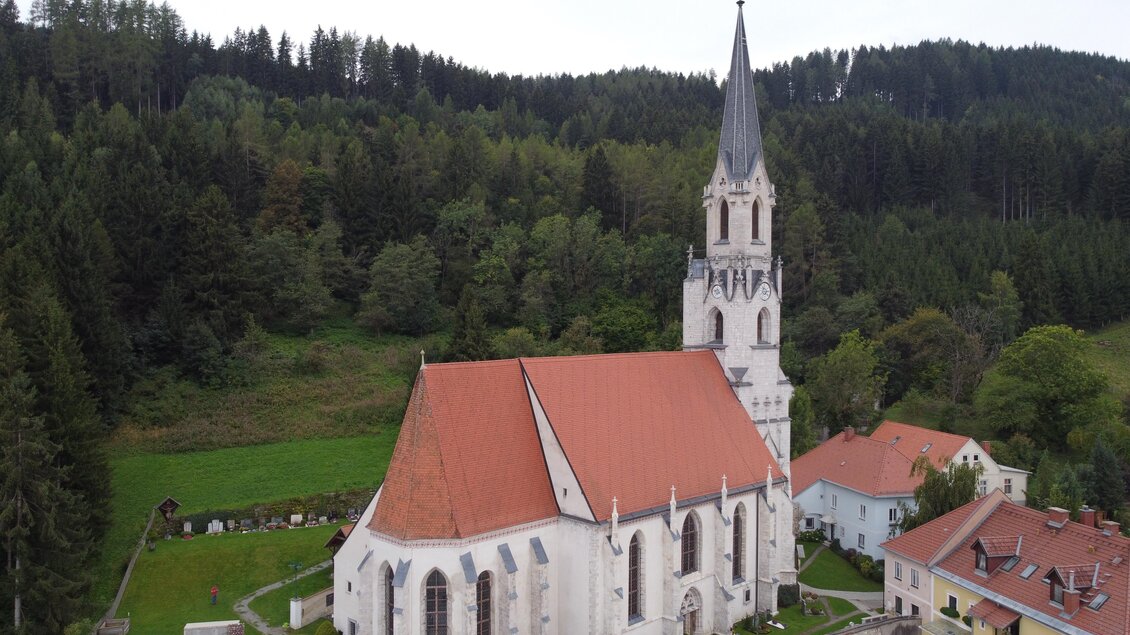 Eine Kirche mit einem hohen Turm und rotem Dach, umgeben von Wäldern und grünen Wiesen. Im Vordergrund sind einige Häuser sichtbar. | © Diazöse Graz Seckau