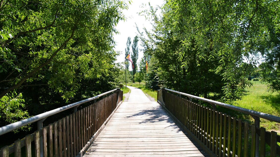 Eine Holzbrücke führt durch eine grüne Landschaft. Umgeben von Bäumen und Licht durchflutet, lädt der Weg zum Erkunden ein. | © Kurkommission Bad Blumau