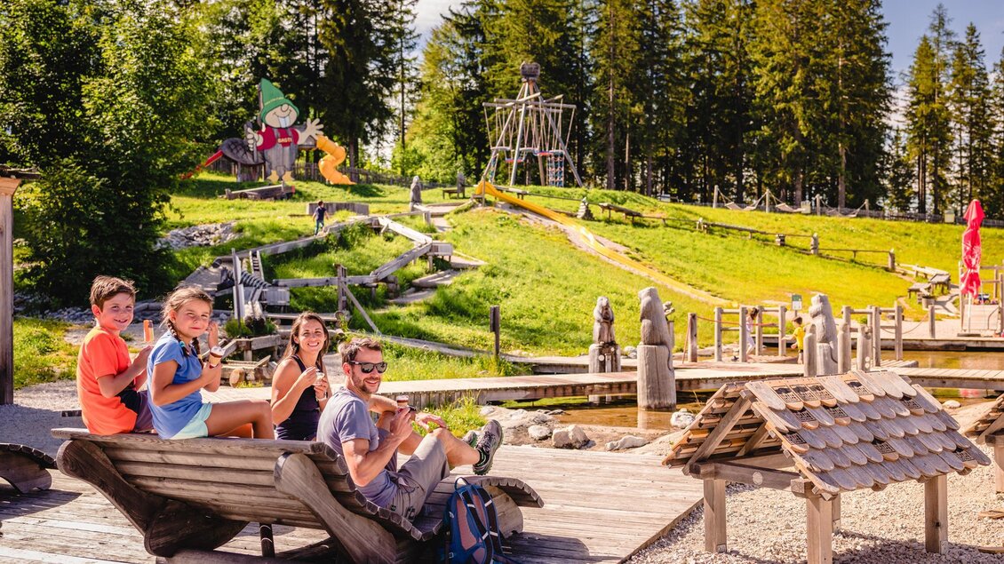Eine Gruppe von vier Kindern sitzt auf einer Bank und genießt die Zeit im Freien. Im Hintergrund sieht man einen Spielplatz mit vielen Aktivitäten und Bäumen. | © Mariazeller Bürgeralpe