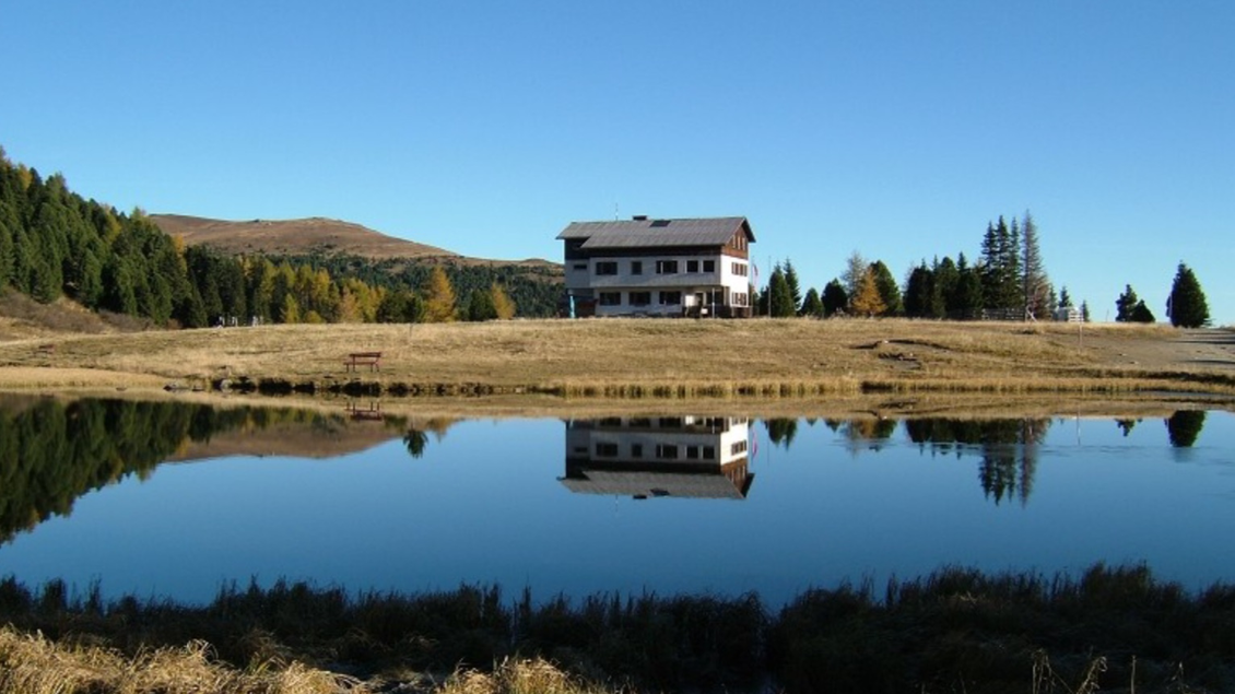 Ein ruhiger See spiegelt ein zweigeschossiges Haus und umgebende Bäume wider. Die Landschaft ist klar und sonnig mit sanften Hügeln im Hintergrund. | © Andreas Wieser