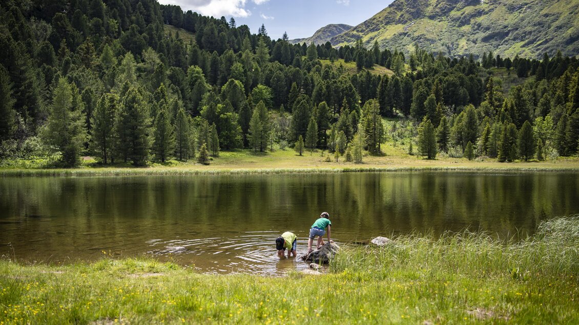 Ein ruhiger See umgeben von hohen Bergen und dichten Wäldern. Zwei Kinder stehen am Ufer und spielen im Wasser. | © Anita Fössl