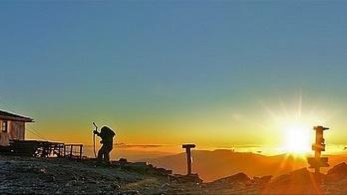 Ein Berghaus steht auf einem Gipfel mit atemberaubendem Sonnenaufgang. Im Vordergrund ist eine Person beim Fotografieren zu sehen. | © Erlebnisregion Murtal
