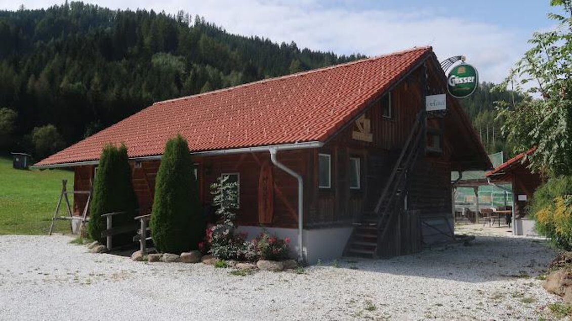 Ein traditionelles Holzhaus mit rotem Dach in einer malerischen Landschaft. Umgeben von Bäumen und einer Wiese, strahlt es ländliche Gemütlichkeit aus. | © s´Hütterl