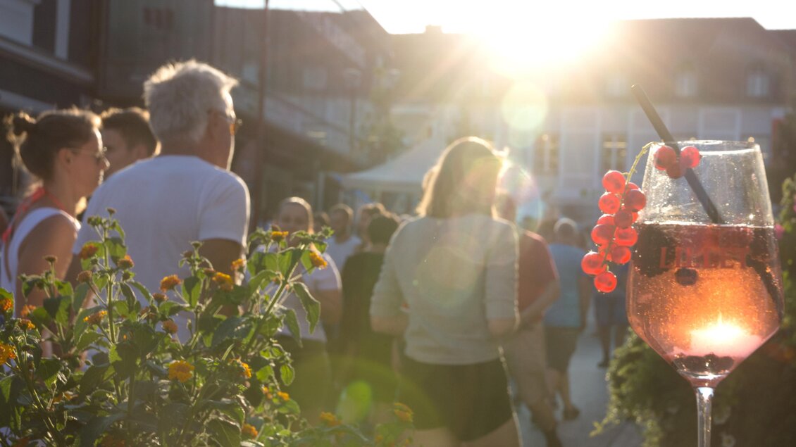 Ein Glas Getränk mit Früchten im Vordergrund und Menschen, die in der Abendsonne spazieren gehen, im Hintergrund. Die Szene vermittelt eine entspannte und fröhliche Atmosphäre. | © Thermen&Vulkanland