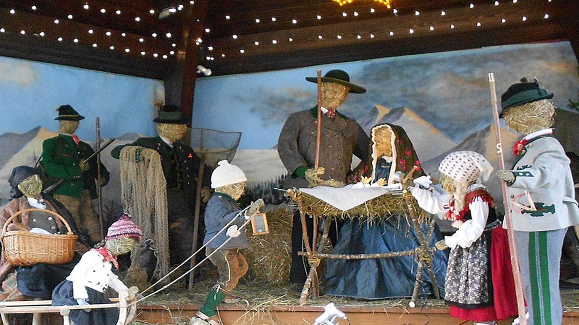 Eine weihnachtliche Krippe mit Strohmännchen in traditioneller Kleidung. Die Figuren sind in einer gemütlichen Umgebung mit festlicher Beleuchtung angeordnet. | © Hildegard Mittermair