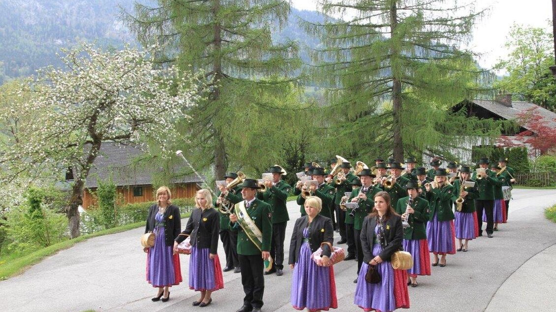 Eine Blaskapelle in traditioneller Tracht steht auf einem Weg, umgeben von Bäumen und Bergen. Die Musiker und Musikerinnen haben ihre Instrumente bereit und lächeln freundlich. | © Hannes Grill