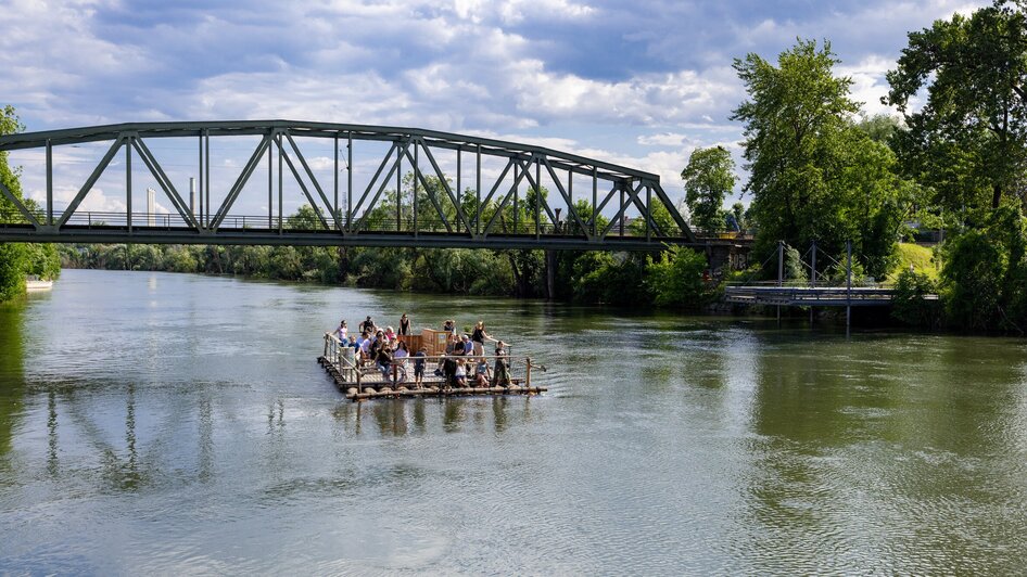 Floß auf der Mur in Graz mit Passagieren und Brücke im Hintergrund. | © Harry Schiffer