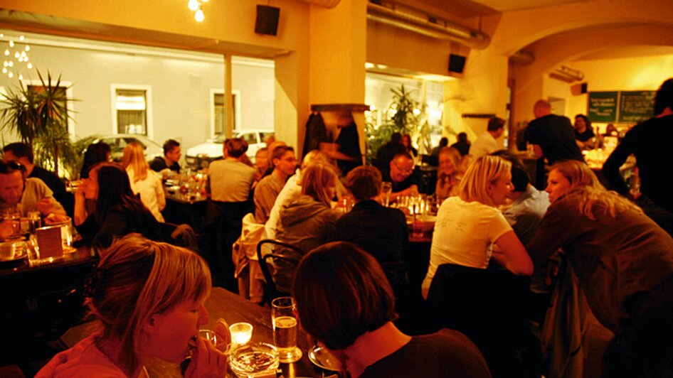 Gemütliches Restaurant mit vielen Gästen, warmem Licht und Pflanzen.