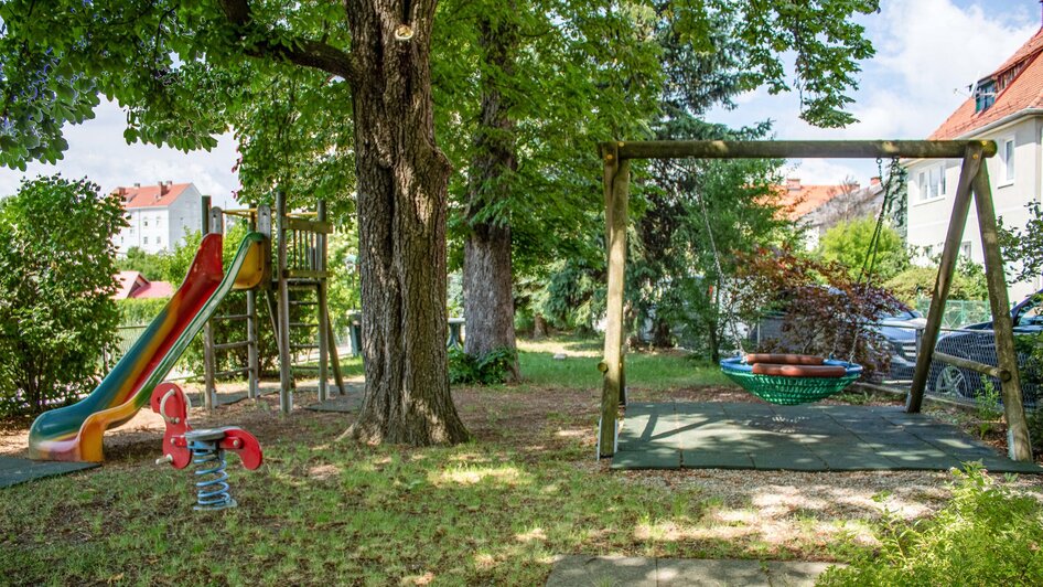Spielplatz mit Rutsche, Schaukel und Wippe in einer grünen Umgebung.