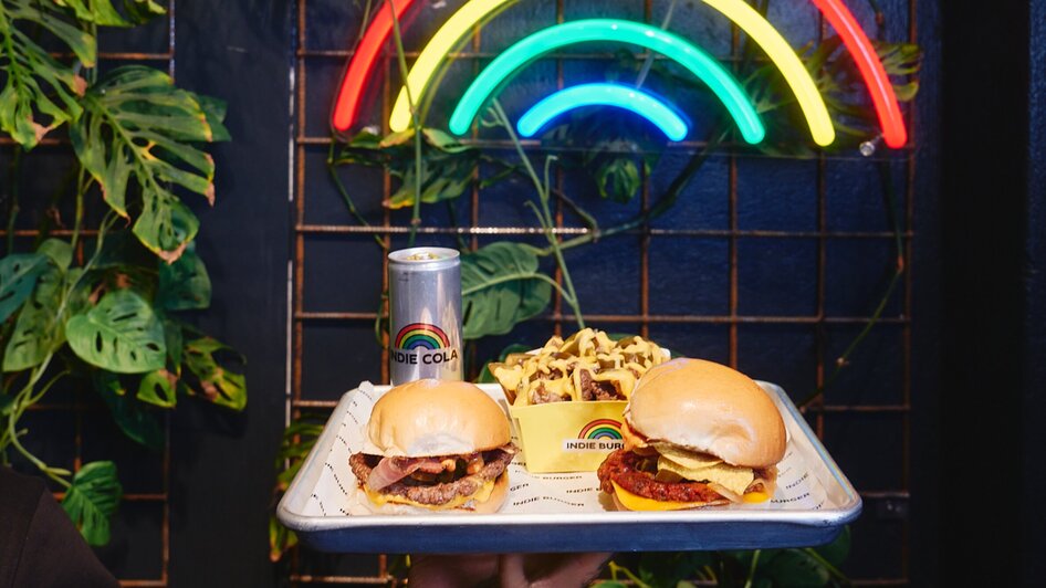 Zwei Burger, Pommes und eine Getränkedose vor einer Neo-Lampe in Form eines Regenbogens.