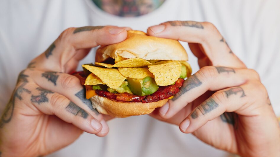 Ein Mann hält einen Hamburger mit Nachos und Avocado-Stücken in seinen Händen.