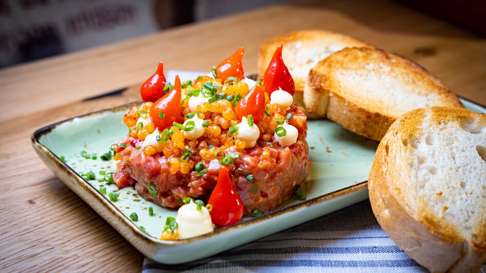 Ein Teller mit Beef Tatar, dekoriert mit Tomaten und Schnittlauch, dazu Weißbrot.