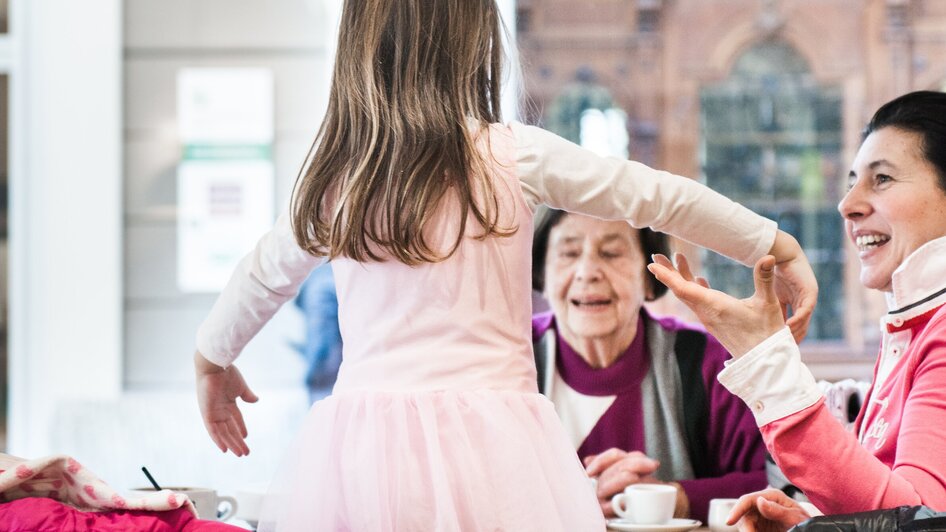 Ein Mädchen in einem rosa Kleid tanzt an einem Tisch mit fröhlichen Erwachsenen in einem Familien-Café in Graz.