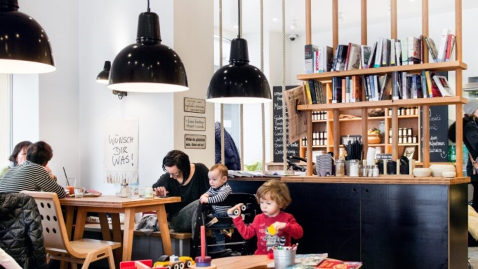 Gemütliches Café mit Bücherregal, Tischen und spielenden Kindern.