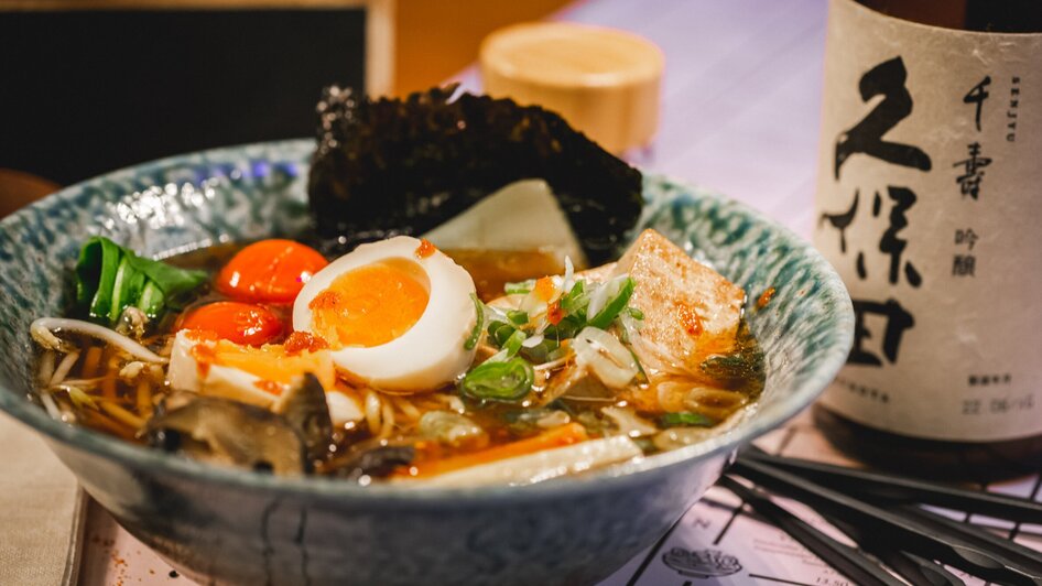Eine Schüssel Ramen mit Ei, Tofu und Gemüse sowie einer Flasche Sake.